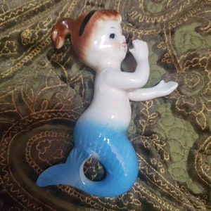May include: A ceramic figurine of a mermaid with a blue tail and a white torso. The mermaid has brown hair with a black headband and is holding her hand to her mouth.