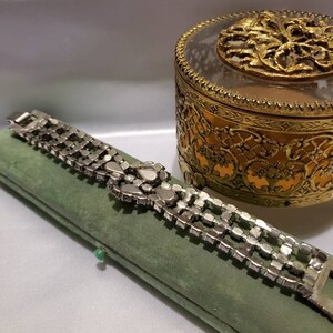 May include: A silver rhinestone bracelet with a large, ornate rhinestone cluster in the center. The bracelet is lying on a green velvet box.