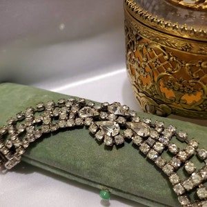 May include: A silver rhinestone bracelet with a large, ornate, teardrop-shaped rhinestone cluster in the center. The bracelet is lying on a green velvet cushion.