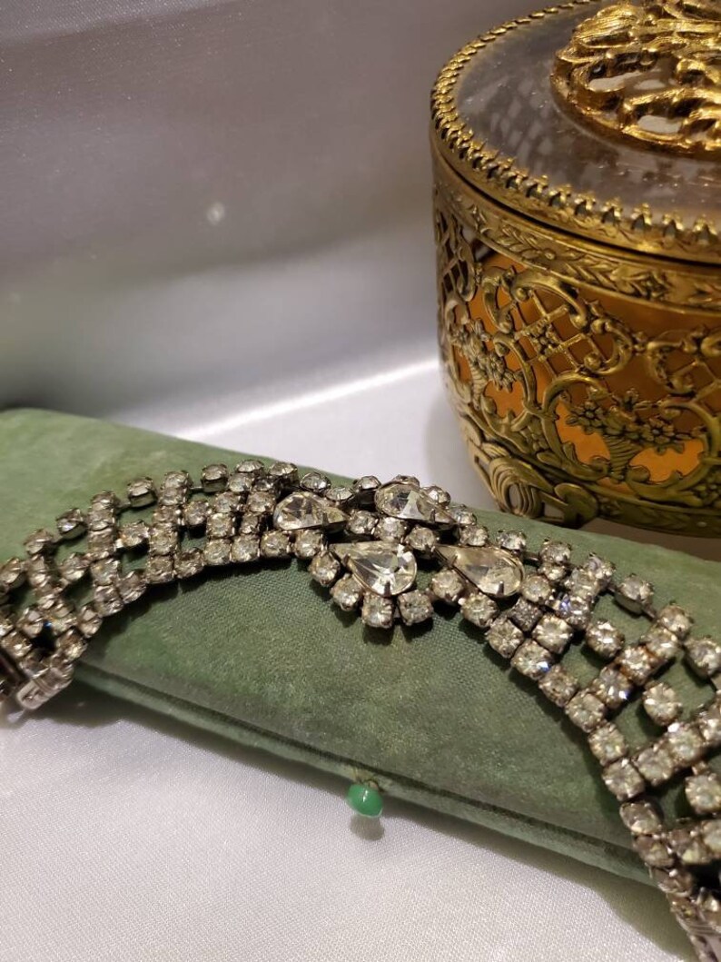 May include: A silver rhinestone bracelet with a large, ornate, teardrop-shaped rhinestone cluster in the center. The bracelet is lying on a green velvet cushion.