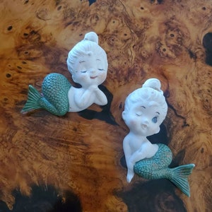 May include: Two porcelain mermaid figurines with green tails and white bodies. The mermaids are sitting with their hands clasped together and have closed eyes.