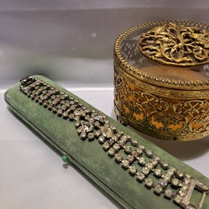 May include: A vintage rhinestone bracelet with a heart-shaped design, displayed in a green velvet box. The bracelet is made of silver tone metal and features clear rhinestones.