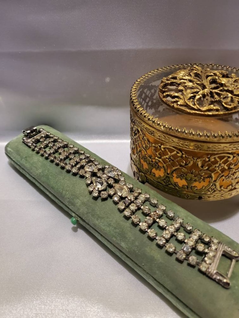 May include: A vintage rhinestone bracelet with a heart-shaped design, displayed in a green velvet box. The bracelet is made of silver tone metal and features clear rhinestones.