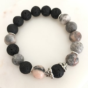 Op de afbeelding: Een armband van zwarte lavasteen en roze en grijze edelstenen met een zilveren bedel. De armband is gemaakt van 8 mm kralen.