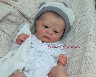 May include: A realistic baby doll with blue eyes, wearing a white onesie with grey stars and a grey and white hat. The doll is lying on a white knitted blanket.