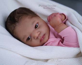 May include: A close-up of a realistic baby doll with brown eyes and dark brown hair, wearing a pink shirt and lying on a white blanket. The doll is made of soft silicone and has a lifelike appearance.