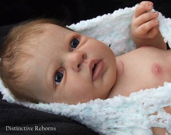 Puede incluir: Un muñeco bebé realista con ojos azules, cabello castaño claro y una tez suave y rosada. El muñeco lleva una manta de ganchillo blanca.