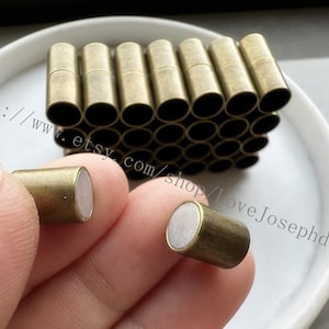 May include: A close-up of a pile of gold-toned metal magnetic clasps. Two clasps are held in a hand, showing the white magnetic surface.