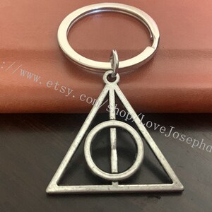 Può includere: Portachiavi d'argento con un ciondolo triangolare che presenta un cerchio con una linea che lo attraversa. Il ciondolo è un simbolo della serie Harry Potter.