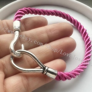 Puede incluir: Un brazalete de cuerda trenzada rosa con un cierre de gancho plateado.