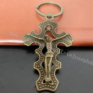 Puede incluir: Un llavero de metal de color bronce con un diseño detallado que presenta una figura de Jesucristo. La cruz tiene un anillo en la parte superior para sujetarlo a un llavero.