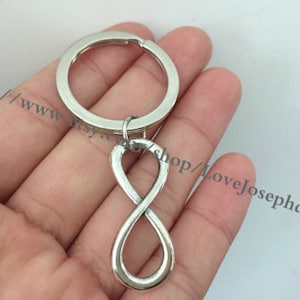 May include: Silver infinity symbol keychain with a silver key ring. The keychain is held in a hand.