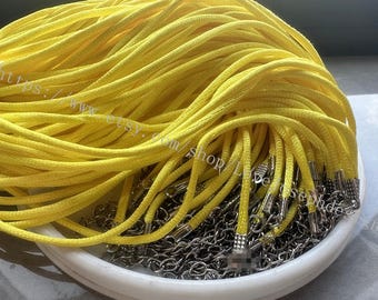 May include: A bundle of bright yellow cord necklaces featuring silver-toned metal clasps and extender chains. The cords are coiled together, with the metallic hardware visible at the ends, resting on a white circular base.