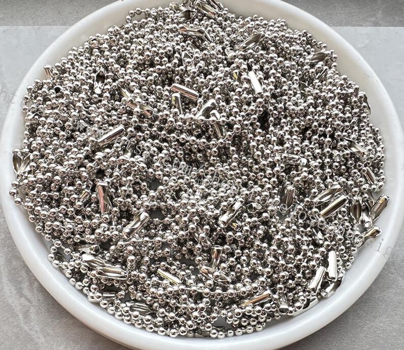 May include: A white bowl filled with a large quantity of silver ball chain. The chain is made of small, round metal beads connected by thin links.