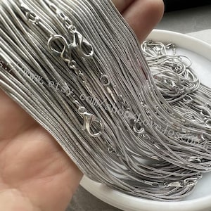 Può includere: Primo piano di un fascio di catene a serpente argentate con chiusure. Le catene sono disposte a ventaglio e sono pronte per essere utilizzate per la creazione di gioielli.