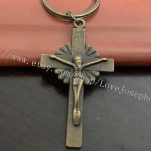 Op de afbeelding: Een sleutelhanger van bronskleurig metaal met een kruis waarop een figuur van Jezus Christus is afgebeeld. Het kruis heeft een decoratief ontwerp rond de randen.