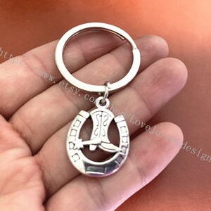 Peut inclure: Porte-clés en argent en forme de fer à cheval avec un charme de botte. Le fer à cheval a un design détaillé et la botte est placée à l'intérieur du fer à cheval.