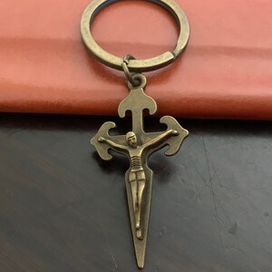 Puede incluir: Un llavero de metal de color bronce con un crucifijo con una figura de Jesucristo en la cruz. El llavero tiene un anillo simple para sujetar las llaves.