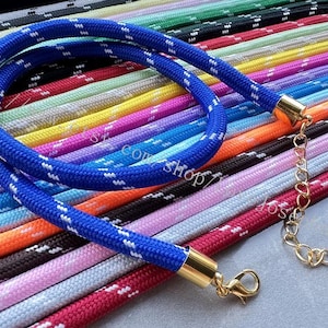 Puede incluir: Un collar de cuerda azul con detalles blancos y cierres metálicos dorados. El collar se muestra con una variedad de collares de cuerda coloridos en el fondo, incluyendo rosa, naranja y negro.