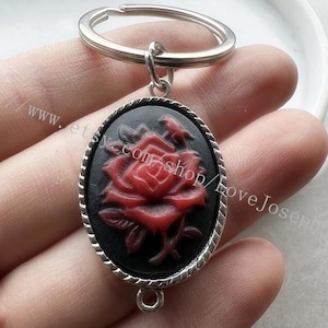 Op de afbeelding: Een ovale sleutelhanger met een zwarte achtergrond en een rood rozenreliëf. Het middelpunt is gevat in een zilverkleurig metalen frame met een gedraaide touwstructuur. Aan de bovenkant zit een zilveren ring en onderaan een klein oogje.