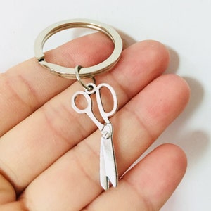 May include: A silver key ring with a silver scissor charm. The keyring is held in a hand.