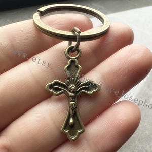 May include: A bronze-coloured cross keyring with a detailed design. The cross has a ring at the top for attaching to a keyring.
