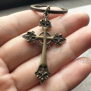 May include: A bronze-coloured cross keyring with intricate details and a ring attached. The cross has a circular design in the centre.