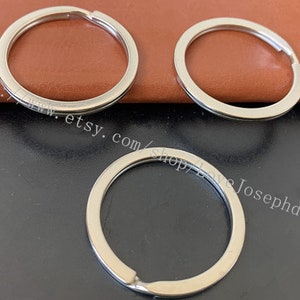 Puede incluir: Tres llaveros de plata, cada uno con forma circular y un acabado liso y pulido. Los llaveros están dispuestos sobre una superficie de cuero marrón.