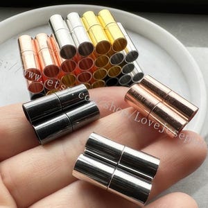 May include: Assortment of magnetic clasps for jewelry making in silver, gold, and rose gold tones. The clasps are arranged in a stack and on a hand.