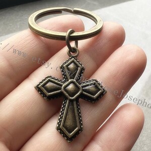 May include: A bronze-colored cross keychain with a detailed design. The cross is attached to a simple metal ring.