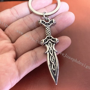 May include: Silver-toned sword keychain with intricate details and a Celtic knot design. The sword has a pointed tip and a rounded hilt.