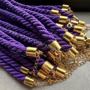 Puede incluir: Un conjunto de cuerdas de poste de cuerda morada con tapas y cadenas de metal dorado. Las cuerdas son gruesas y retorcidas, con un color vibrante y saturado. Las tapas y cadenas doradas añaden un toque de elegancia. La imagen está bien iluminada.
