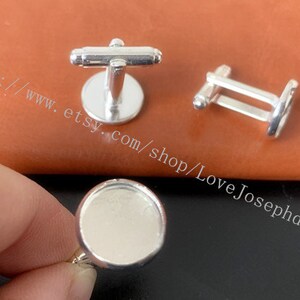 May include: Silver cufflinks with a round, blank, silver-toned cameo setting. The cufflinks are on a brown leather surface.