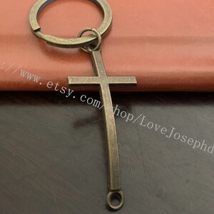 May include: A bronze cross keychain with a simple design. The cross is attached to a ring with a loop at the bottom.