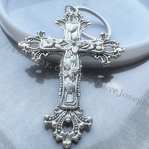 May include: Silver-toned cross keychain with intricate floral details. The cross is attached to a silver-toned ring.