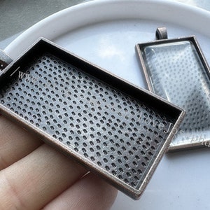 May include: Two antique silver-toned metal picture frames with glass inserts. The frames are rectangular and have a textured surface. The frames are designed to be used as pendants.
