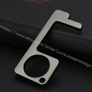 May include: A silver-toned key-shaped tool with a loop on the end. It has a small hook on one side and a larger hook on the other side. The tool is designed to help people open doors and press buttons without touching surfaces.