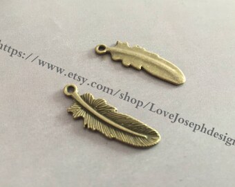 Puede incluir: Dos abalorios de metal en forma de pluma de color dorado antiguo con un bucle en la parte superior para sujetar a joyas o manualidades.