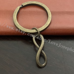 May include: A bronze key chain with an infinity symbol charm. The key chain is on a brown leather surface.