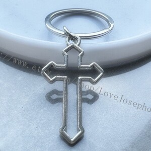 May include: Silver-toned metal keychain featuring a cross design. The cross is open and has a simple, elegant design.
