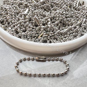 May include: A close-up of a silver ball chain, a type of jewelry chain made of small metal balls connected by links. The chain is displayed in a white bowl, with a smaller section of the chain lying on a white surface.
