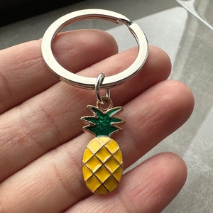 Puede incluir: Un llavero de plata con un dije de piña. La piña es amarilla con hojas verdes y un tallo plateado.