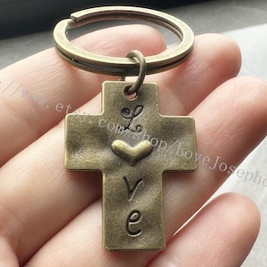 May include: A bronze-colored cross keychain with a heart and the word "love" engraved on it. The keychain is attached to a silver key ring.