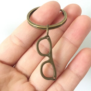 Könnte beinhalten: Ein Schlüsselanhänger aus Bronze mit einem Brillen-Anhänger. Die Brille ist aus Metall und hat ein schlichtes Design.