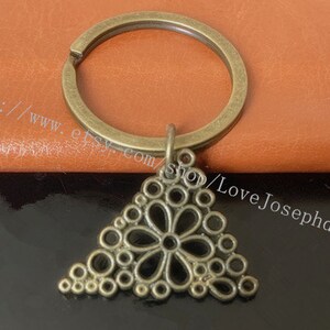 Può includere: Un portachiavi in bronzo con un ciondolo triangolare che presenta un design floreale decorativo. Il ciondolo è realizzato con un'intricata lavorazione in metallo con un motivo circolare.