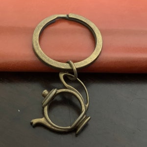 Puede incluir: Un llavero de metal dorado con un dije de tetera. El dije de tetera tiene un cuerpo redondo y un pico. El llavero está unido a un anillo plateado.