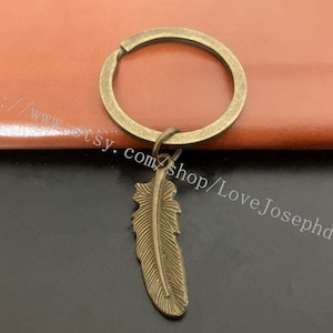 以下が含まれることがあります： 羽のペンダントが付いたブロンズ色のキーホルダー。 羽にはエッチングされた線が施され、キーホルダーのリングは円形です。 背景は茶色と黒の混合です。