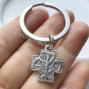May include: Silver key chain with a cross charm featuring the Chi Rho symbol. The cross is detailed with a textured surface.