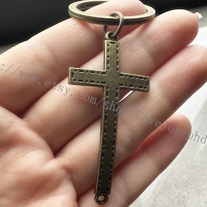 May include: A bronze-colored cross keychain with a detailed design. The cross has a ring at the top for attaching to a key ring.