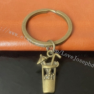 May include: A bronze keychain with a charm of a cocktail glass with a straw and an umbrella. The keychain is attached to a bronze key ring.